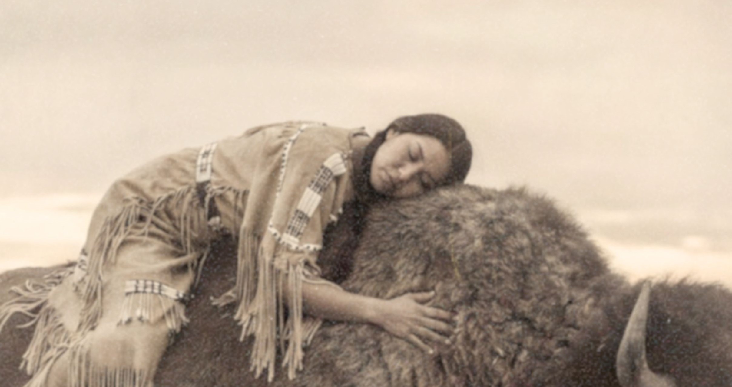 Close-up detail of a sepia-toned fine art print showing a Native American woman with braided hair and fringed traditional clothing lying with her eyes closed across the broad back of a bison, her arms wrapped gently around the animal as swirling prairie mist surrounds them and a massive UFO looms silently through hazy clouds above, revealing the rich aged texture and warm vintage tones of this surreal Western art print.