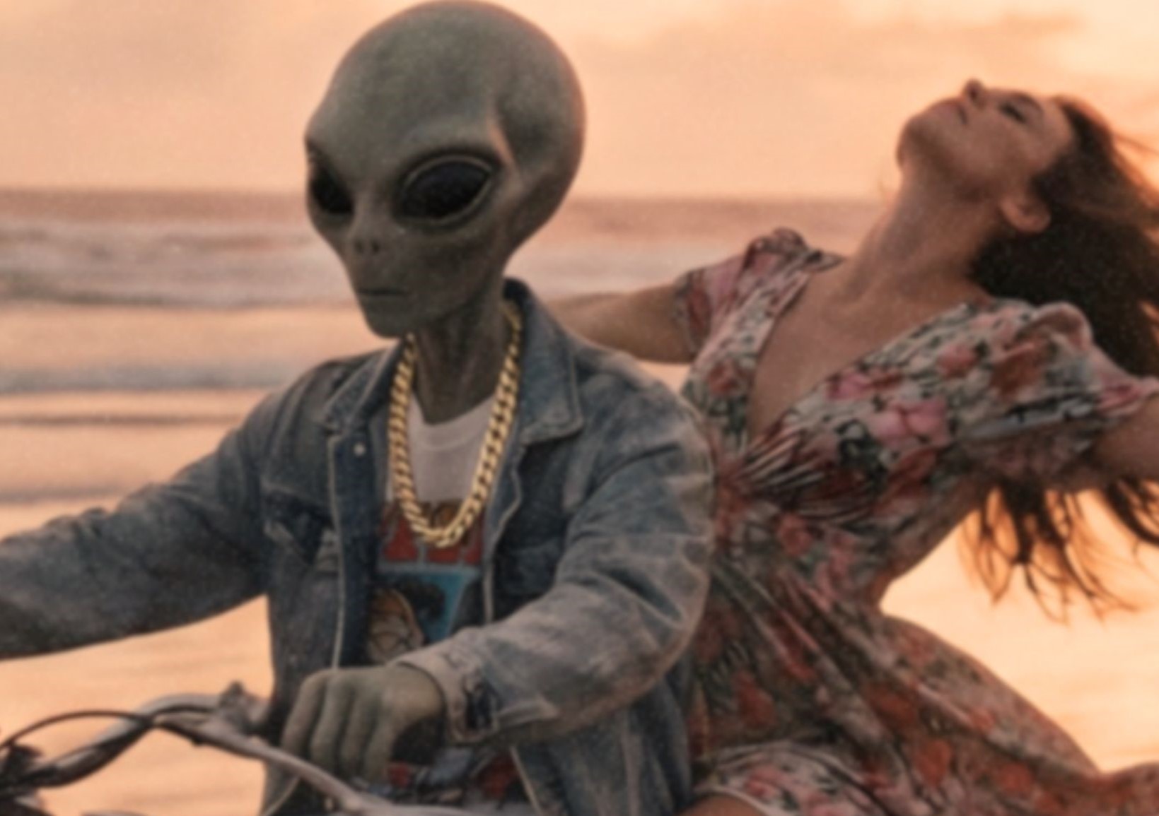 Intense warm-toned close-up of a grey alien in a denim jacket and gold chain gripping a vintage motorbike handlebar on a glowing sunset beach with a UFO looming in golden clouds above – unframed retro alien portrait poster, ufoposter.com