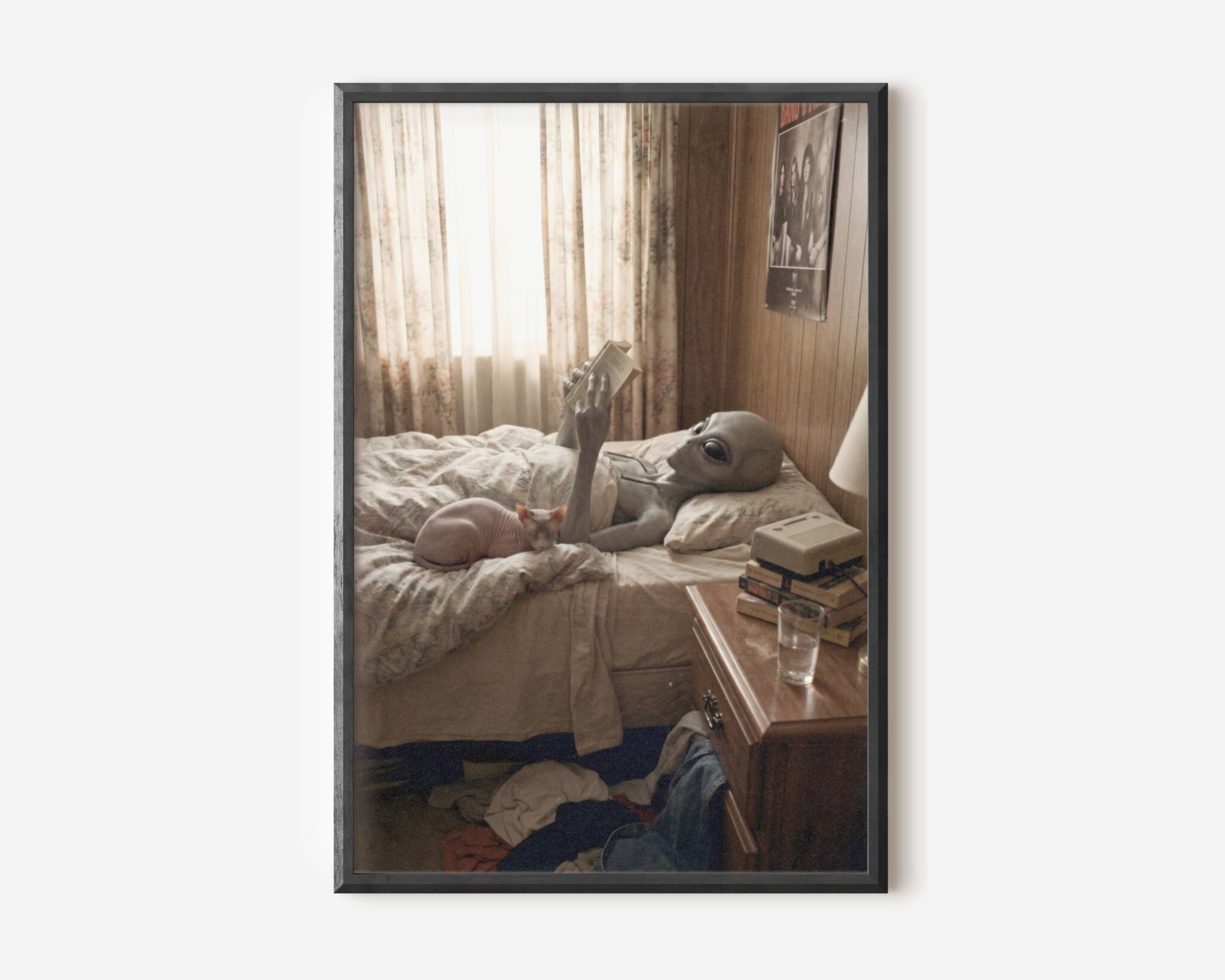 Premium funny retro sci-fi poster of a grey alien lying on a rumpled bed reading a book beside a sphynx cat in a warm wood-panelled bedroom with VHS tapes, floral curtains and a glass of water on the nightstand – unframed alien art poster, ufoposter.com