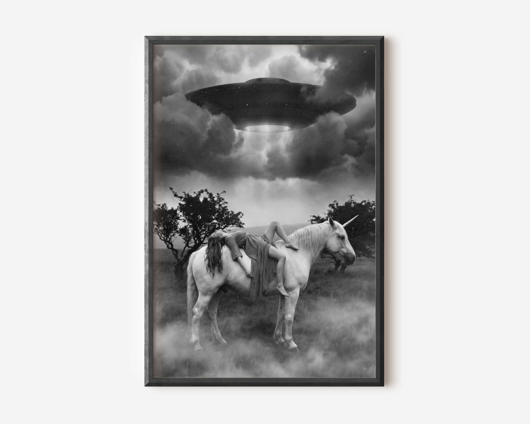 Black and white fine art print of a woman draped across the back of a white unicorn standing in a misty field, as a large UFO hovers above emitting a beam of light through dramatic storm clouds. A surreal and cinematic sci-fi wall art print printed on premium matte paper.