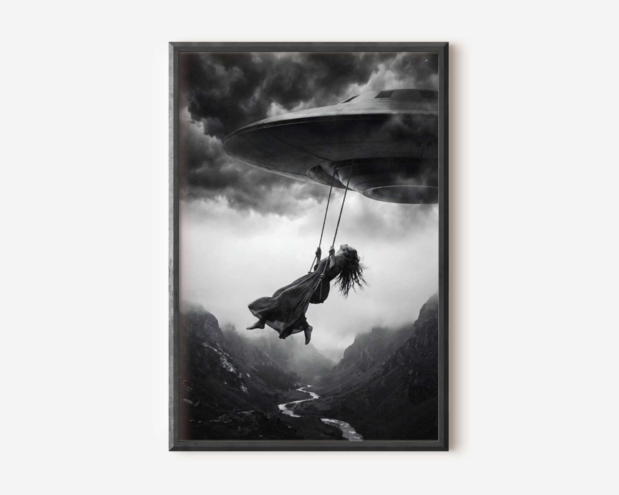Black and white fine art print of a woman in a flowing dress swinging from a rope attached to a UFO spacecraft, suspended high above a dramatic mountain canyon under a stormy sky. A surreal and cinematic sci-fi wall art print printed on premium matte paper.