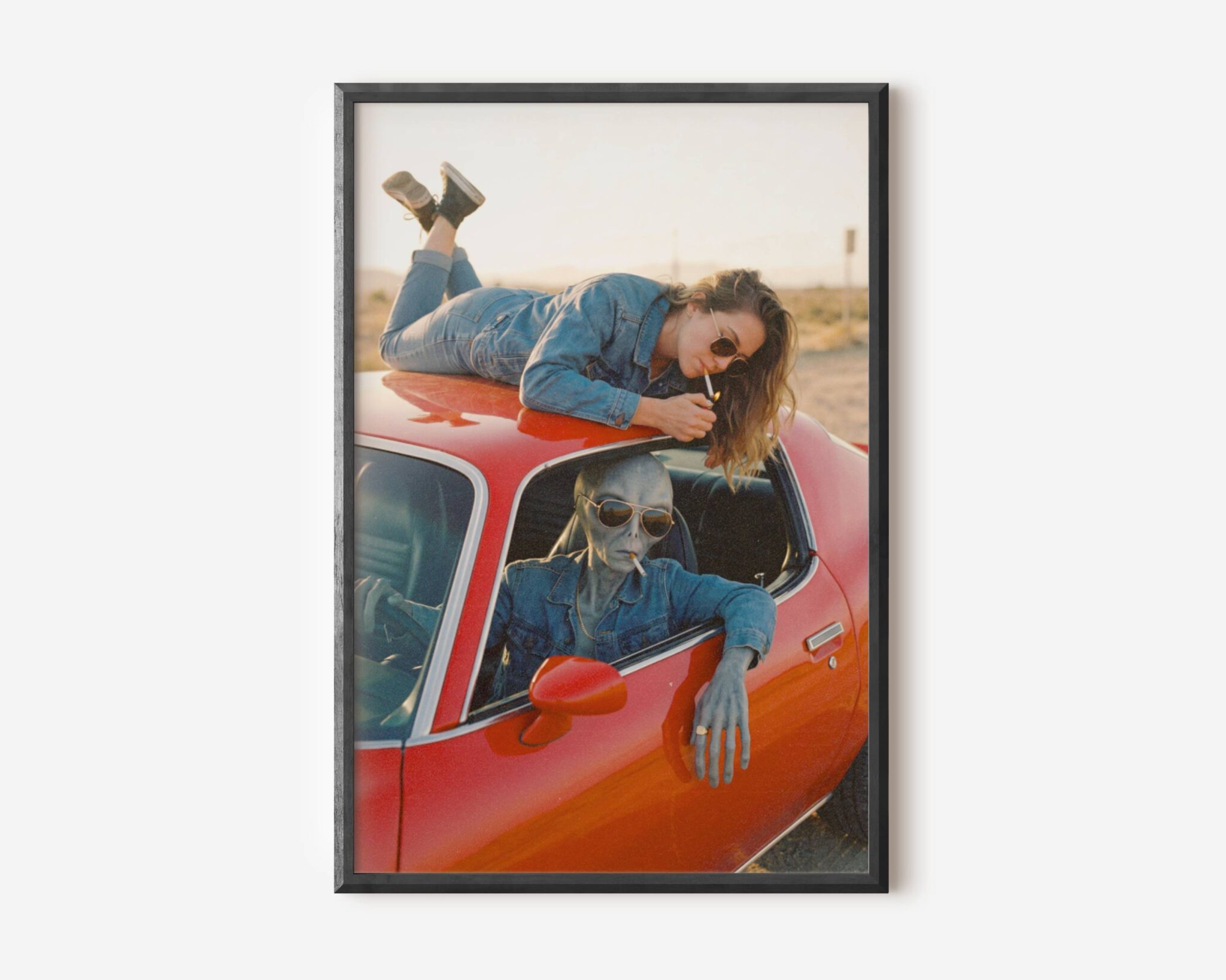 Premium retro sci-fi poster of a grey alien in aviator shades and denim jacket driving a vintage red car through a desert landscape with a woman lounging on the roof lighting a cigarette – unframed alien art poster, ufoposter.com
