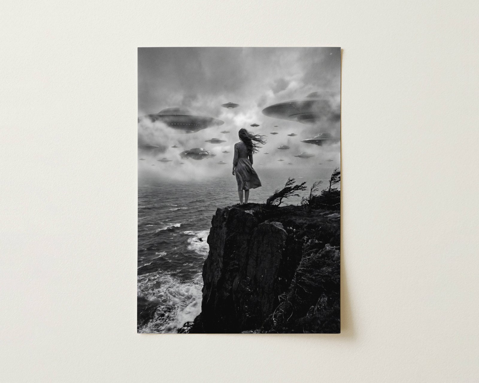 Black and white fine art print photographed flat as a sheet of paper, showing the full print quality and matte texture. The image depicts a lone woman with hair blowing in the wind facing a fleet of flying saucers emerging from swirling fog, capturing the cinematic mood and fine detail of this sci-fi wall art print.