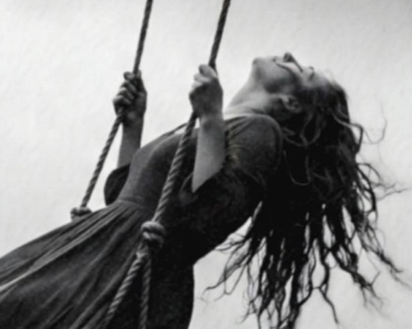 Close-up detail of a black and white art print showing a woman with wild flowing hair gripping a rope swing attached to the underside of a UFO spacecraft, her dress and hair caught in the wind as she hangs above a deep mountain canyon.