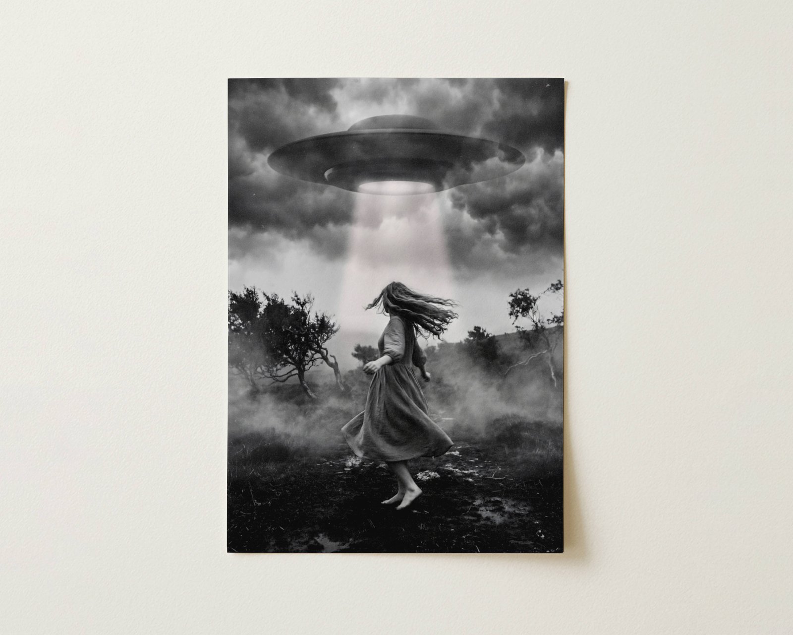 Black and white fine art print photographed flat as a sheet of paper, showing a barefoot woman rising off a misty moorland ground beneath a large UFO beaming light through turbulent storm clouds, surrounded by twisted dark trees and swirling fog. The flat lay highlights the rich tonal contrast and fine detail of this surreal cinematic art poster.