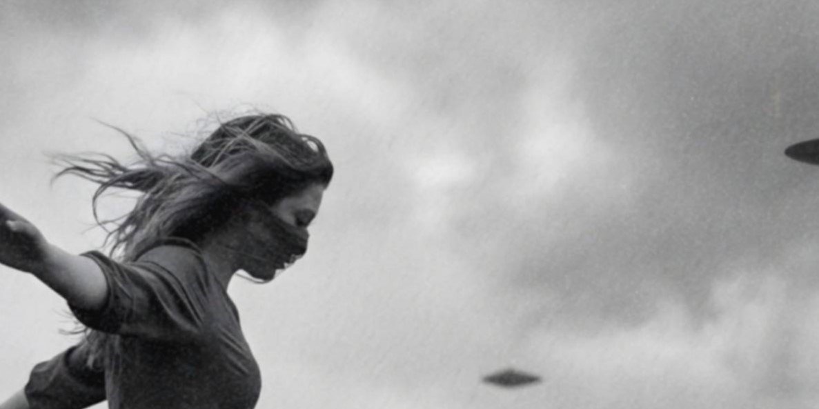 Close-up detail of a black and white art print showing a woman in a dress balancing on the edge of a rocky cliff with her arms outstretched, one foot stepping onto a small hovering UFO as her hair blows in the wind and flying saucers circle in the cloudy sky behind her.