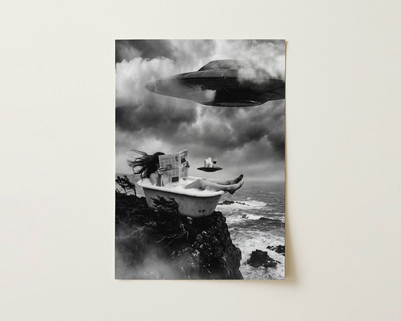 Black and white fine art print photographed flat as a sheet of paper, showing a woman soaking in a clawfoot bath on a sea cliff reading a newspaper while a large UFO descends through dramatic storm clouds and a small UFO acts as a floating drinks tray beside her. The flat lay highlights the rich tonal contrast and fine detail of this surreal cinematic art poster.