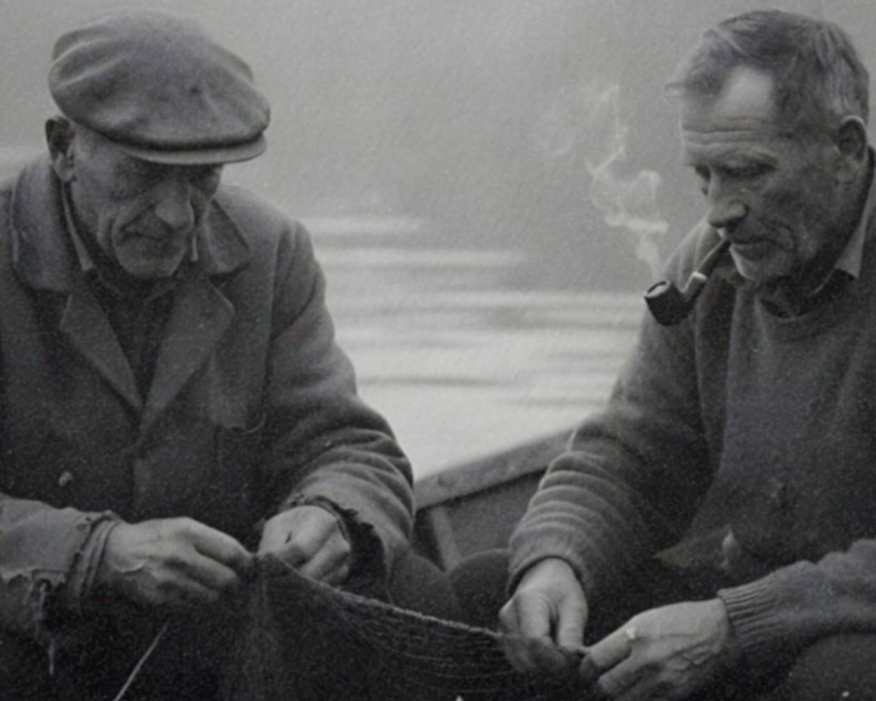 Intense vintage close-up of two old fishermen absorbed in mending their nets in a rowboat, a looming UFO silently materialising from dense fog on the water behind them – unframed dark alien portrait poster, ufoposter.com
