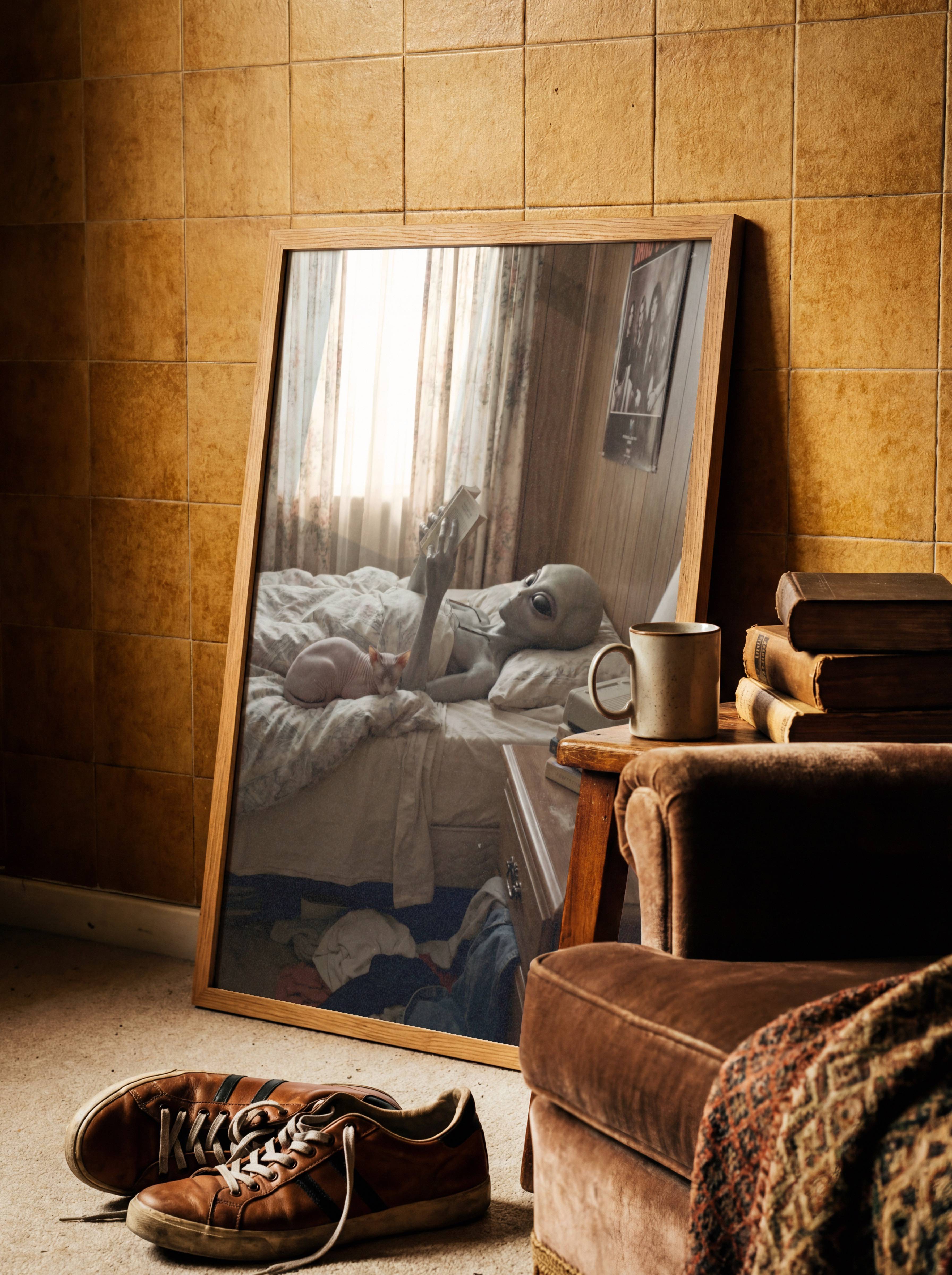 Deeply charming warm vintage retro portrait of a grey alien having an entirely perfect lazy afternoon — rumpled duvet, sphynx cat, a good book, a glass of water and absolutely no intention of saving the world today – unframed retro sci-fi art poster, ufoposter.com