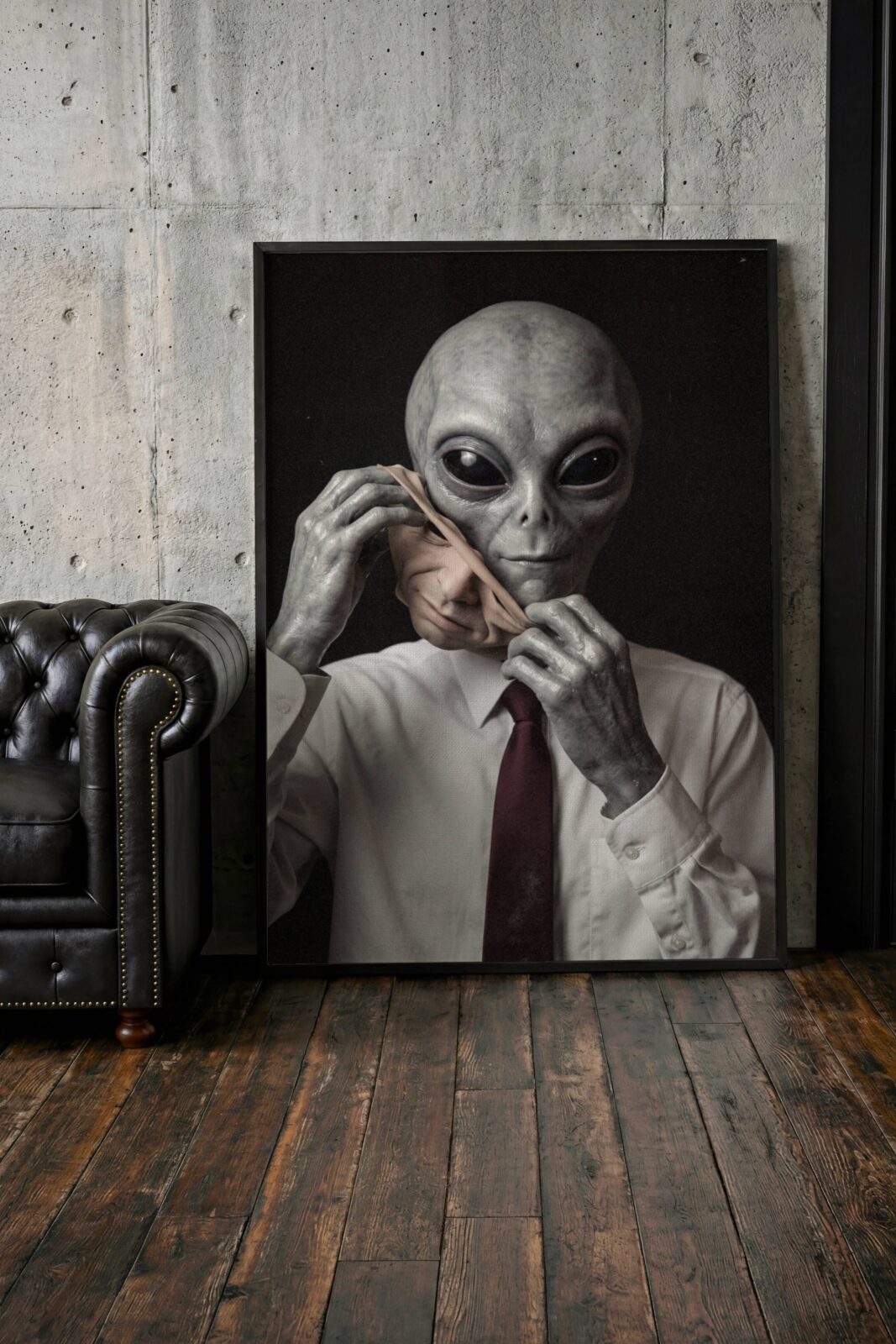 Unsettling dark artwork of a grey alien in a business shirt and red tie methodically removing a human face mask, suggesting a world where aliens walk undetected among us – unframed dark alien wall poster, ufoposter.com