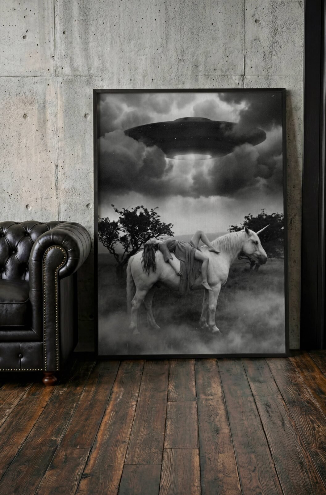 Black and white fine art print of a woman draped across a white unicorn standing in swirling mist beneath a large hovering UFO and turbulent storm clouds, displayed in a study or home library, adding a visually powerful and imaginative surreal quality to an intellectual interior.