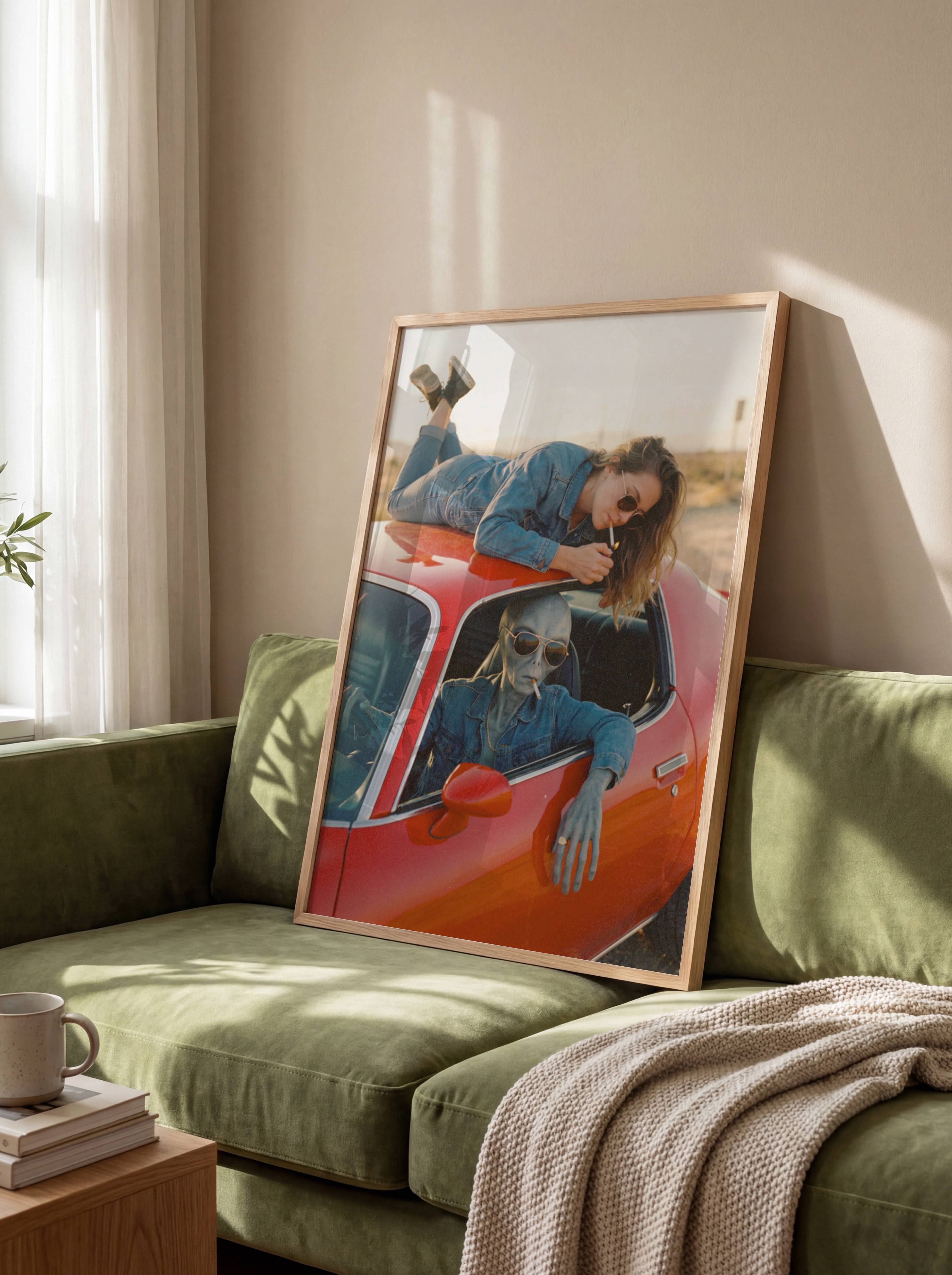 Bold retro sci-fi portrait of a grey alien in a denim jacket and aviator sunglasses sharing a desert road trip with a relaxed woman draped across the roof of a vintage red car – unframed dark retro alien wall poster, ufoposter.com