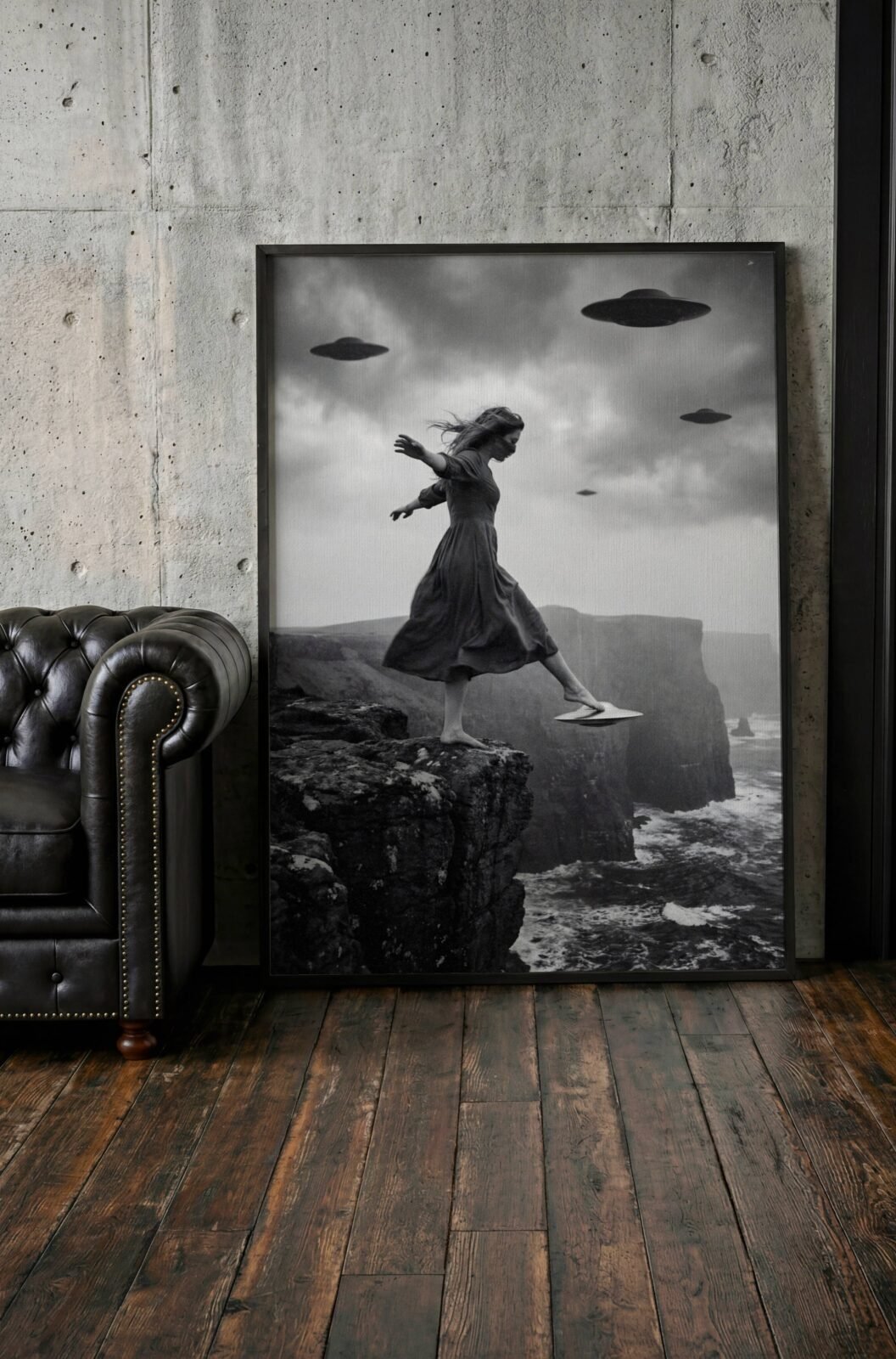 Black and white fine art print of a woman stepping from a rocky sea cliff onto a small hovering UFO as flying saucers circle in a dramatic stormy sky above a wild coastline, leaning against a wall in a lifestyle flat lay setting, shown unframed to highlight the premium matte paper quality and rich tonal contrast of this cinematic surreal wall art poster.