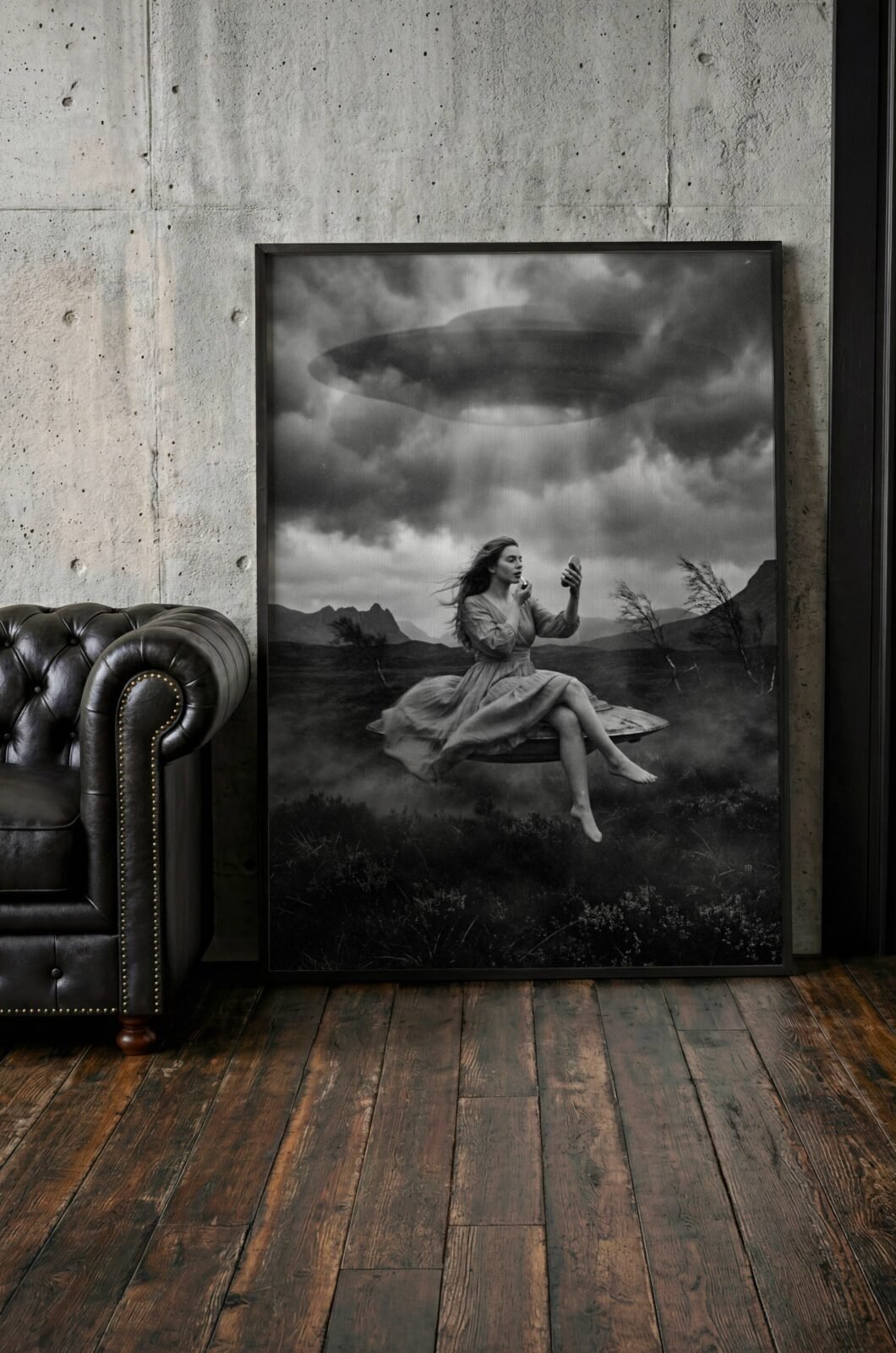 Monochrome surreal art print of a woman floating on a levitating platform over a moody landscape while applying makeup beneath a large UFO in a stormy sky, displayed in a Scandinavian-style interior, adding cinematic depth and dry wit to a minimal space.