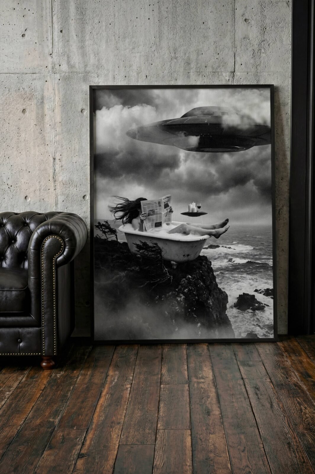 Black and white fine art print of a woman reclining in a clawfoot bath on a rocky coastal cliff, reading a newspaper with complete calm as a large UFO looms through storm clouds and a small saucer hovers with candles and wine beside her, leaning against a wall in a lifestyle flat lay setting, shown unframed to highlight the premium matte paper quality and rich tonal contrast of this cinematic surreal wall art poster.