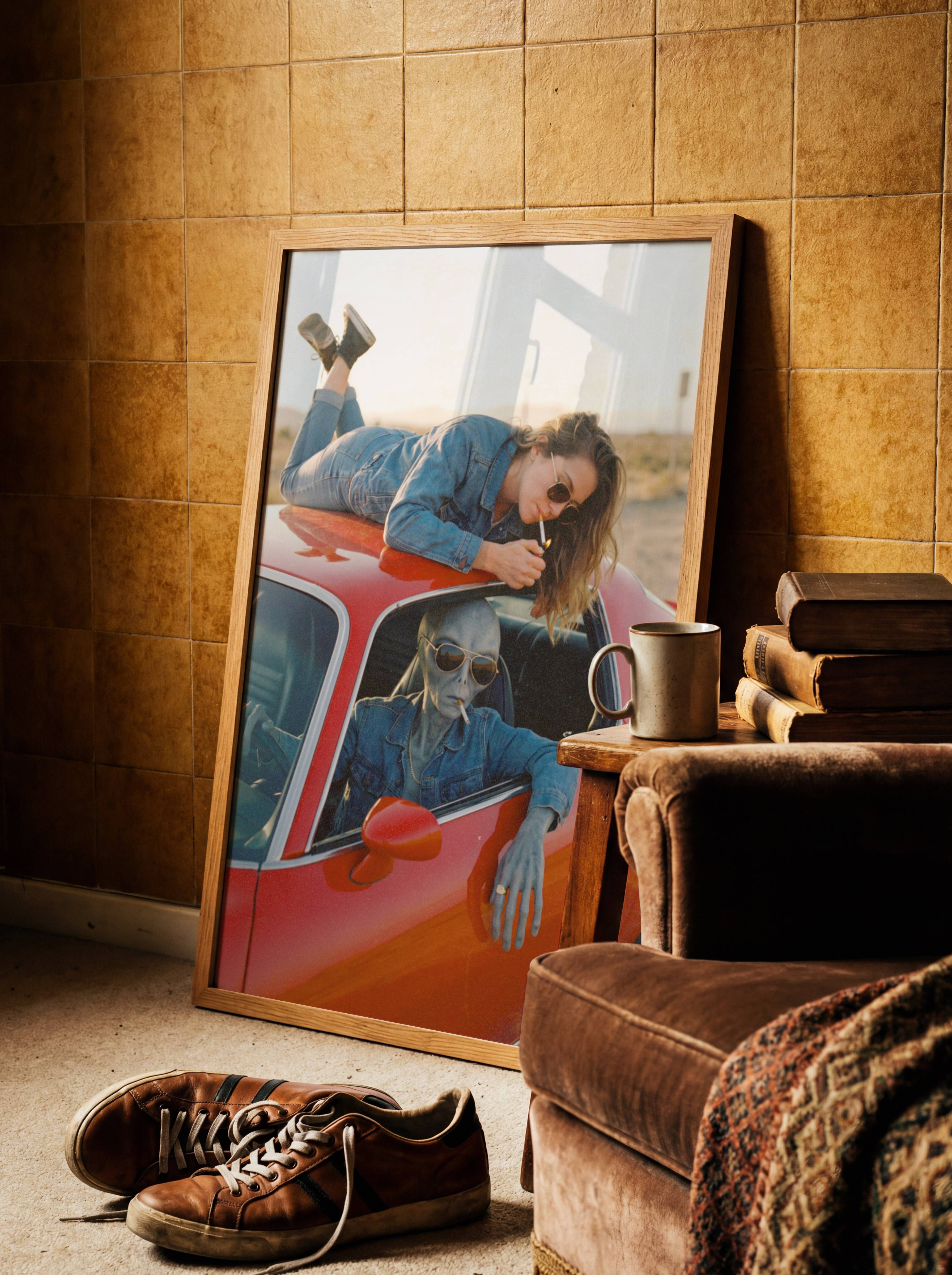 Angled retro shot of a grey alien in aviator shades leaning coolly from a vintage red car window as a woman in denim lies across the roof lighting a cigarette in golden desert light – unframed retro sci-fi wall poster, ufoposter.com
