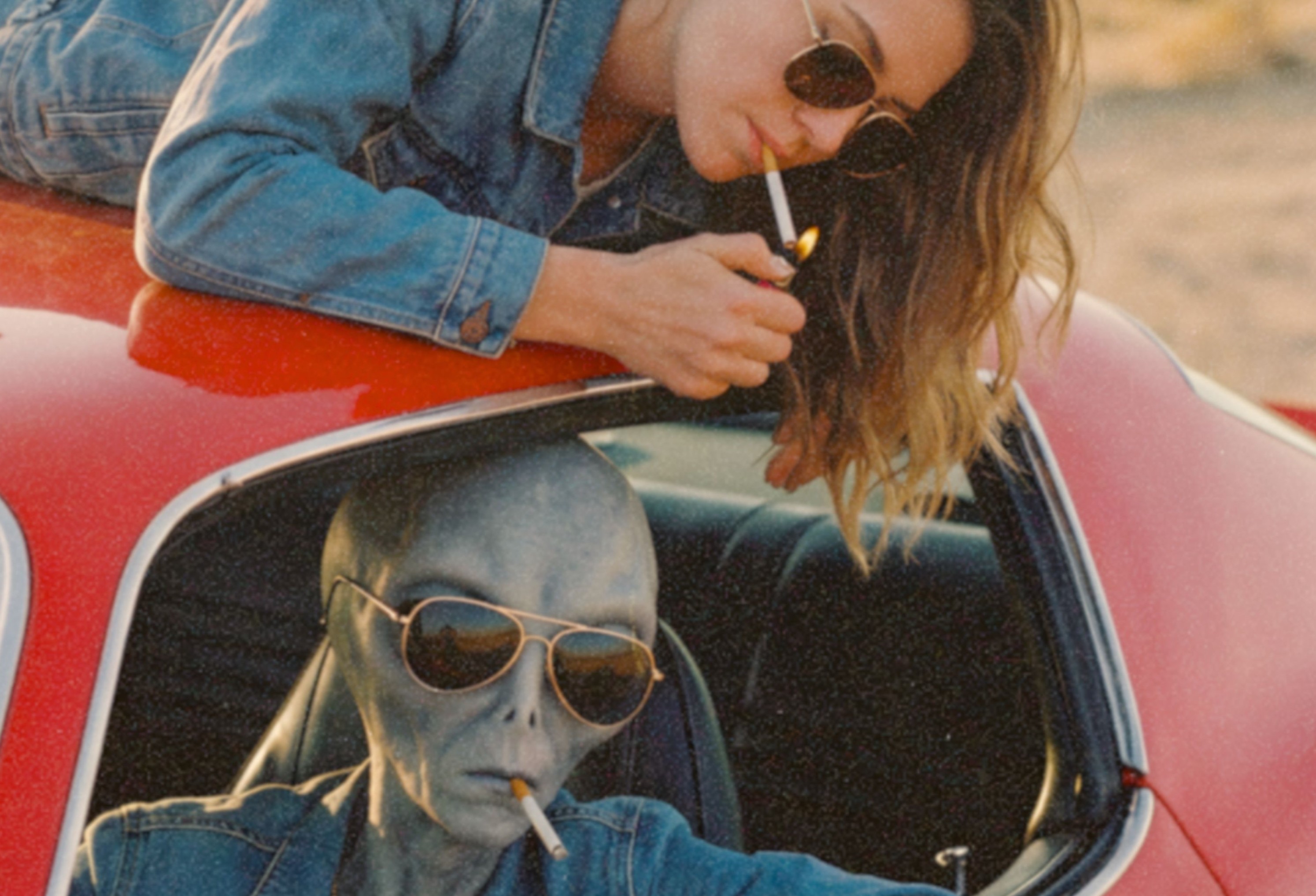 Intense close-up of a grey alien in a denim jacket and aviator sunglasses with a cigarette between its lips leaning out of a classic red car window in warm desert light – unframed retro alien portrait poster, ufoposter.com