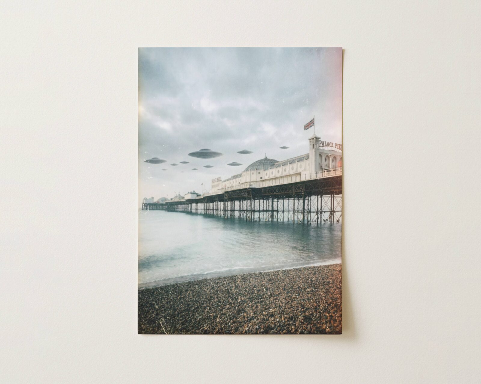 Flat print soft atmospheric retro artwork of a fleet of UFOs above Brighton Palace Pier in a pale grey winter sky, shingle beach and calm grey-green sea below — high quality unframed flat poster print, ufoposter.com