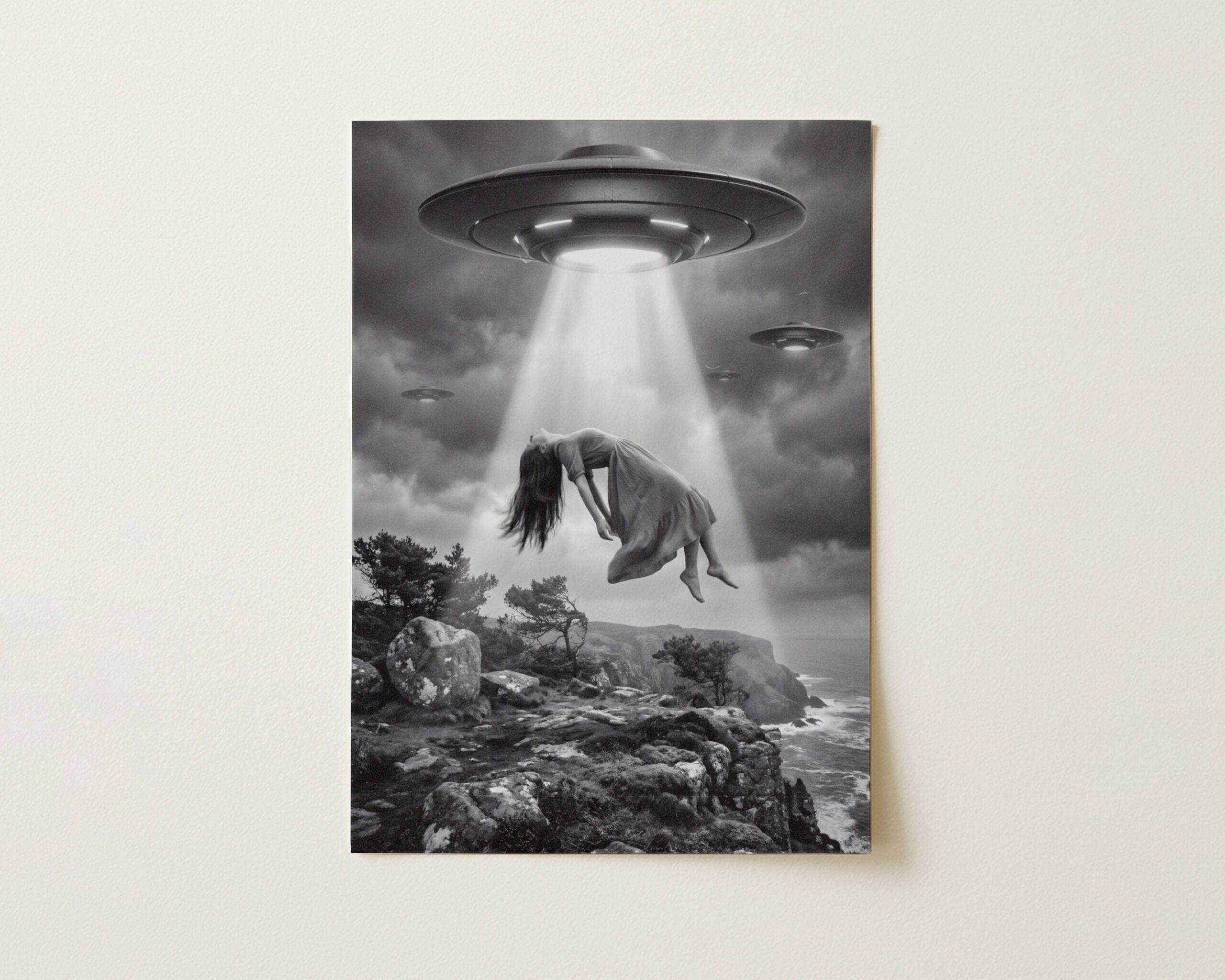 Flat print atmospheric black and white surreal artwork of a woman floating limp in a blinding UFO beam above a dramatic rocky coastal clifftop with two more spacecraft in heavy storm clouds and a grey churning sea below — high quality unframed flat poster print, ufoposter.com