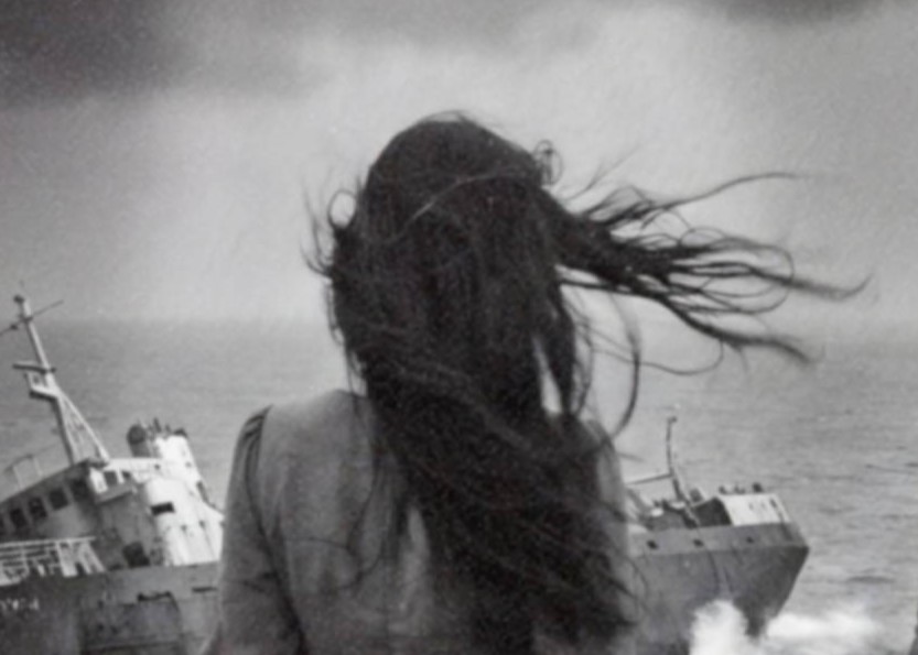 Intense black and white close-up of a barefoot windswept woman's back as she walks toward a stormy coastline, a shipwreck listing in crashing waves, a glowing UFO beam above in dark clouds and a unicorn visible on a clifftop rock arch to the right – unframed dark surreal detail poster, ufoposter.com